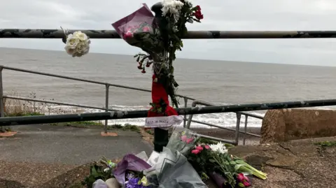 The picture shows a memorial set up along a coastal walkway overlooking the sea. A metal railing runs across the scene, and attached to it are bouquets of flowers, including white roses and mixed arrangements of red and white blooms. More flowers are laid on the ground, wrapped in paper and plastic, with colours ranging from purple and pink to white and green. 