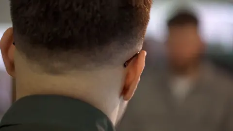 A man the BBC is calling Ishaan to protect his identity is looking at the reporter, who us ahead but blurred out. Ishaan is facing away so his face cannot be seen. He has short hair and the arms of glasses are curved round the back of his ears.