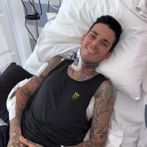 Tai Woffinden/Instagram A man with dark hair and tattoo-covered arms smiles into the camera. He is lying in a hospital bed surrounded by medical equipment. He is wearing a black vest with a green logo.