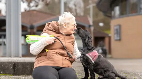 Sue Davis, who has short grey hair and glasses, is wearing a brown gilet and black leggings. She is sat on a kerb with her nose to the nose of her black cocker spaniel Arlo.
