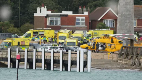 Eddie Mitchell Multiple ambulances and and air ambulance can be seen behind a handful of parked cars at the Shioreham waterfront. The sea can be seen in the foreground, with breakwaters. Behind the ambulances are houses. Several people can be made out amound the emergency vehicles
