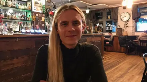 BBC Alex Garden sitting in the Greenbank Pub. They have long blonde hair and are wearing a dark top.