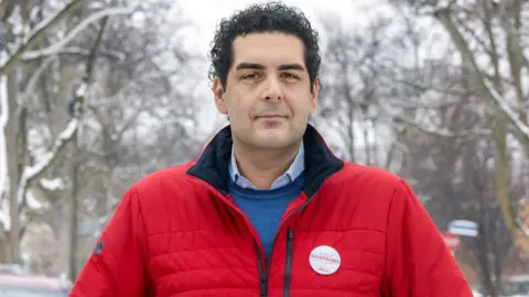 Getty Images Vince Gasparro, he is looking at the camera, he has black hair and is wearing a blue jumper and shirt with a red jacket.