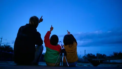 Getty Images A father and two children sit on a wooden deck at dusk, facing a deep blue evening sky. All three are pointing upward at faint stars, and a small telescope on a tripod stands beside them as they stargaze together.