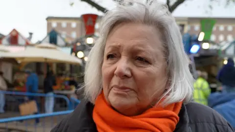 Qays Najm/BBC Sarah Langshaw mira a la cámara. Tiene el pelo gris hasta los hombros y viste una bufanda naranja y una chaqueta negra. Está parado frente a un mercado.