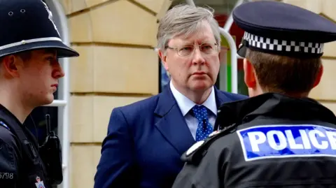 Roger Hirst is wearing a navy suit and blue tie while talking with two male uniformed police officers. He has grey hair and is wearing glasses.