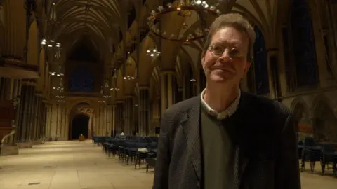A tall man stands inside a large cathedral. He has bushy brown hair and wears glasses, a brown jacket, white shirt and olive green jumper. The cathedral has sand-coloured paving stones and stone walls and pillars.