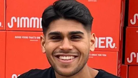 A man of South Asian heritage smiles at the camera. He is wearing a black T-shirt. Behind him is a stack of red food delivery boxes.