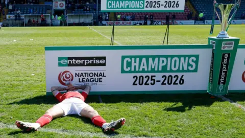 PA Media A man in a red and white football kit lying on a pitch in front of a stand that says Champions 2023-2026