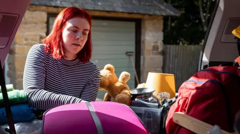 Getty A female student with straight red shoulder-length hair is packing a pink suitcase into the boot of a car. She wears a black and white stripey top and is arranging various objects, including a teddy, a set of towels and a yellow lampshade, in the boot. Behind her is a garage with a green door.