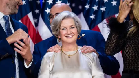 Getty Images Susie Wiles, who has grey hair and is wearing a shiny white dress, smiles as Chris LaCivita clasps her shoulders from behind