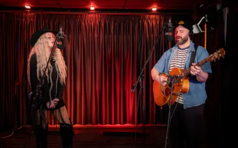 Janet Devlin is wearing all black and a black hat. Her guitarist is wearing a denim jacket over a stripy white and blue top with black trousers and a cap. They are in a studio with red curtains and lights. 