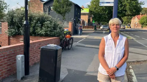 LDRS Paula Widdowson - a woman with short blonde hair, wearing shorts and a white shirt. She is standing in front of a bus stop on a residential street