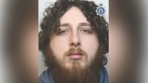 A police mugshot of a man with long brown curly hair and a brown beard. He is wearing a blue-coloured hooded top.