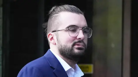 Eddie Mitchell A man with glasses and brown hair tied up. He has brown facial hair and is wearing a dark blue suit jacket and white shirt.