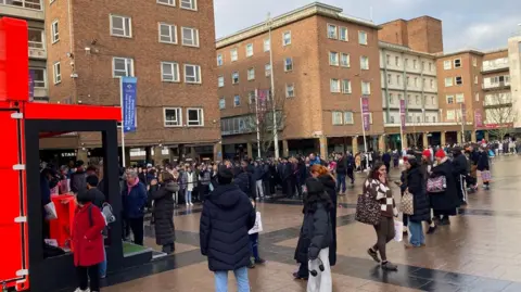 People are standing in a city centre square surrounded by shops. They are wearing coats and queuing at a red stand that is selling football shirts.