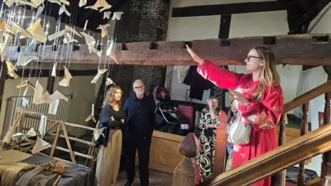 Olha Barvynka Olha is seen standing on some stars with one of her arms outstreched towards an installation which shows what appears to be paper planes hanging from the ceiling. She is wearing a long-sleeved pink dress. There are three other people standing in the room