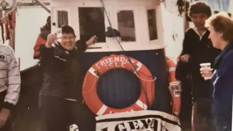 Fiona Brown Moira is standing in front of a boat smiling and holding up a drink.