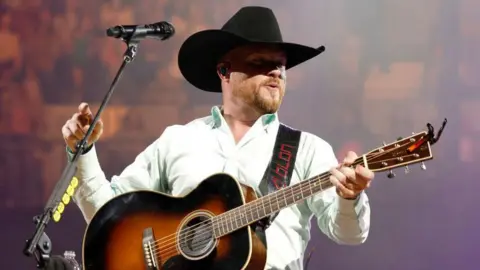 Cody Johnson is wearing a black cowboy hat, a white shirt with the top buttons open, and he is playing a black and brown guitar with fans visible behind him.