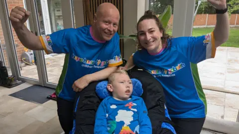The couple are wearing blue hospice T'shirts and stand behind their son sitting in a special chair with their arms raised.