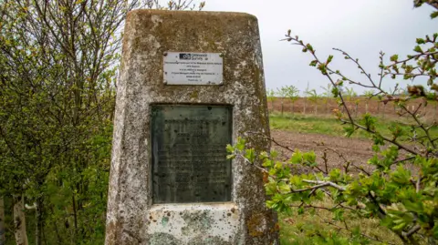 McCoy Wynne Photography The Cold Ashby Trig Pillar in Northamptonshire. It has a plaque and sits in between small trees, with a field behind it.