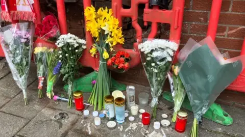 Several bunches of flowers are lined up along a red brick wall. On the ground are several red and white candles. 