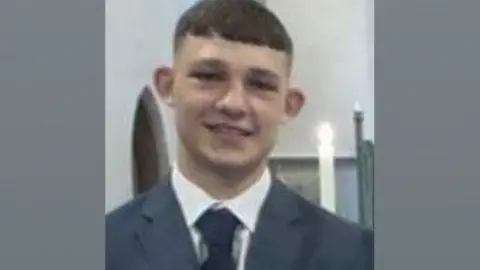 Nojukas Korzeniauskas,  a young man wearing a grey jacket over a white shirt and dark tie. He has short dark hair cut in a short fringe over his forehead and shaved at the sides. Behind him can be seen a lit candle. 