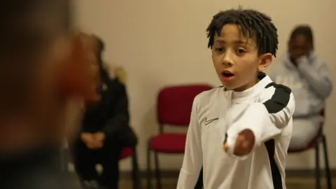 Eidan is wearing a black and white top and pointing accusingly