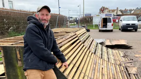 Guy Campbell/BBC Carl Hurr standing next to wooden slats of a a skatepark ramp
