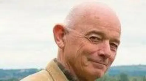 Ted Stevens from Hucclecote Parish Council is wearing a tan jacket over a dark shirt, looking directly at the camera with a slight smile. 