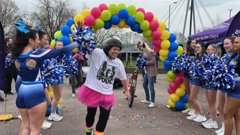 A woman wearing a T-shirt of the band The Police, a cycling helmet, a pink tutu and black leggings is smiling as she passes underneath a balloon arch. On either side of her are cheerleaders in blue leotards and holding blue and silver pom-poms.