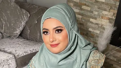 Ambreen Nawaz Ambreen Nawaz is wearing a light green-blue hijab with gold trim and is sitting in a living room with a silver sofa behind her. There is a wall behind her covered in small brick-like tiles.