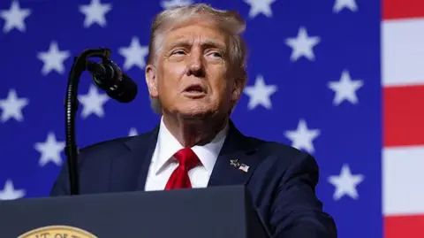 President Donald Trump speaks to an audience while standing in front of a brightly lit background with the American flag 