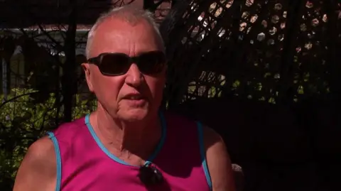 Man with grey hair and glasses. He's wearing a pink vest.