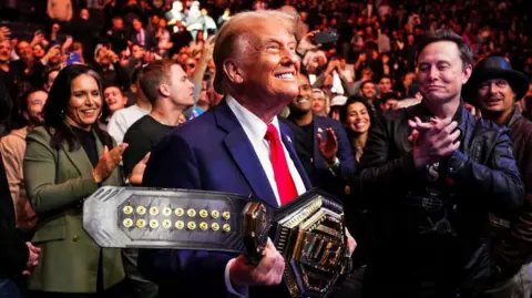 Getty Images Donald Trump grins while holding the UFC belt presented to him by UFC fighter Jon Jones. He is surrounded by a crowd that includes Elon Musk and Tulsi Gabbard. 