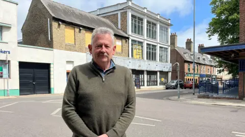 Kim Leonard, who has grey hair and moustache, is wearing a green quarter-zip jumper and standing across the street from 33-39 St James Street in King's Lynn. Some of the nine windows are filled with signs advertising the garage to the rear
