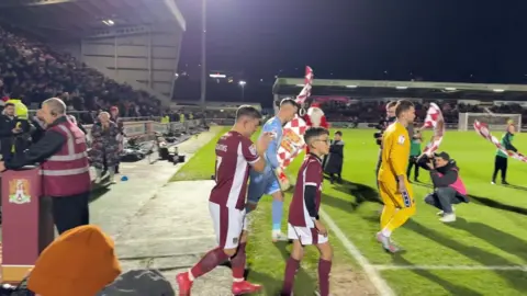 Ezra and captain Sam walk out onto the pitch along with members of Northampton Town and the other team, AFC Wimbledon. Stewards or mascots wave chequered red and white flags, along with a person dressed as Santa. You can see two of the four sides of the stadium which is packed with crowds. It is night time and the pitch is floodlit.