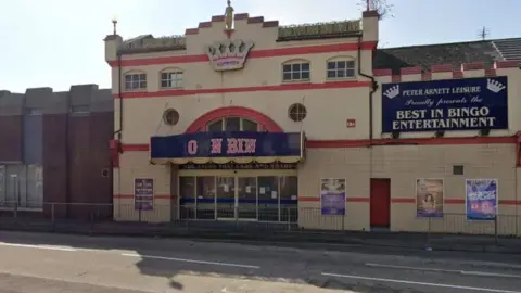 Exterior view of a bingo hall on a town street. The cream-coloured building has red trim, a crown-shaped decoration above the entrance and large signs advertising bingo entertainment. A blue canopy covers the main doors, with posters displayed on the front of the building. The road and pavement in front are empty in bright daylight.