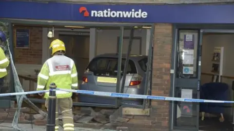 Trevor Porter A firefighter stands in front of Nationwide, behind a blue and white striped cordon. The window of the bank is completely smashed, with bricks and glass strewn about. A silver Honda Jazz is visible inside the branch.