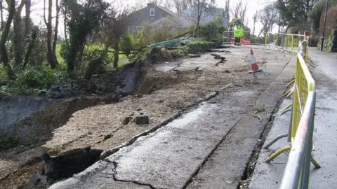 Isle of Wight Council Undercliff Drive, with large cracks and holes in the tarmac. The area is fenced off and a policeman is on duty in the background.