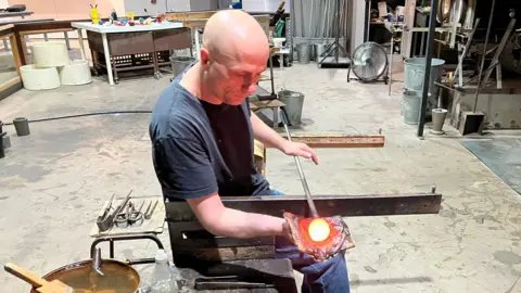 A man demonstrates how molten orange glass expands into a globe shape at the tip of a metal rod. He is wearing a dark shirt and is in a room with various metal tools placed around.