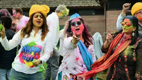 Three women covered in coloured powder are dancing. One has a white tshirt with rainbow writing saying 'Happy Holi'.