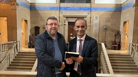 Peterborough First Mohammed Farooq and Chris Harper jointly hold a black book, while they stand in front of a grand staircase. The words "The City of Peterborough" are above a door behind them. Farooq is dressed in a dark suit, and Harper is wearing a blue polo shirt and navy jacket.