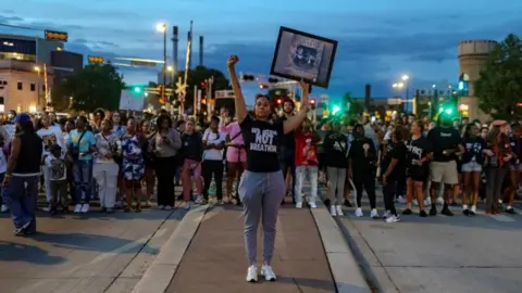 A woman in a black shirt that reads "we been not breathing" holds her fist and her hand in the air while other protesters stand in the background