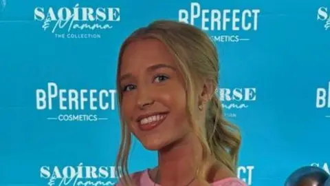 A woman with blonde hair wearing a pink top stands smiling in front of a blue wall with different brand names written in while. She is standing beside grey and blue balloons.