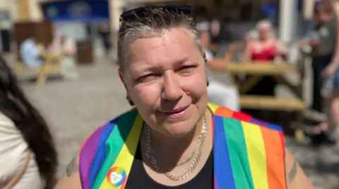 Sam Read/BBC Allison is squinting slightly in the sun while facing the camera. She is wearing a black top, chains around her neck and a rainbow sleeveless jacket. She has short hair and is smiling.