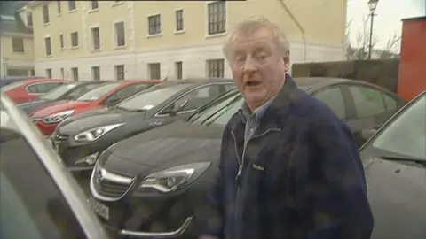 RTÉ A man with grey hard stands in a car park. He is wearing a blue and white shirt and a navy blue jacket.