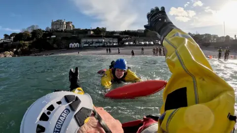 Caroline Pellow swimming towards the casualty in the water. She is pushing a red buoy towards him. The casualty has his arms in the air.