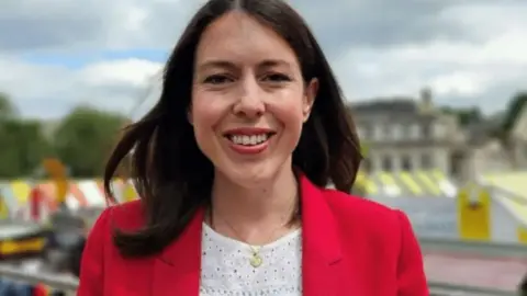 Paul Moseley/BBC MP Alice Macdonald in front of Norwich Market. She is wearing a red jacket over a white top. She has shoulder-length dark hair and is smiling at the camera. The colourful striped roofs of the market stalls are behind her. 