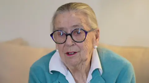 Jamie Niblock/BBC Shelley Cowlin, a woman who is sitting down on a cream sofa. She is not looking directly at the camera and is wearing a white blouse with a teal cardigan over the top. Her hair is tied back and she is wearing a pair of circular framed glasses.